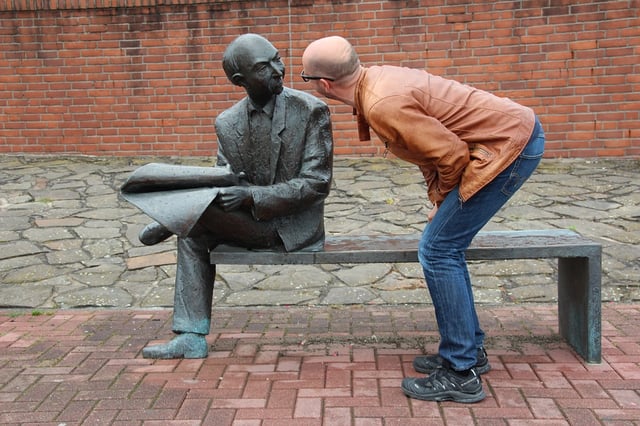 Stock photo of sculpture of a sitting man observed by a bystander.